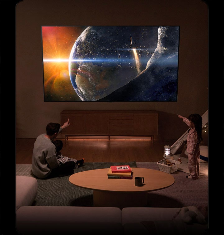 Una familia sentada en el suelo de una sala de estar poco iluminada frente a una mesa pequeña mirando un LG TV montado en la pared que muestra la Tierra desde el espacio.