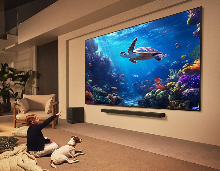 A huge LG QNED TV and LG Soundbar is mounted on the wall. A little girl and her puppy are watching an underwater scene on the TV. The sheer size of the TV makes the content feel even more immersive.