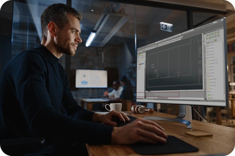 A man seated at a desk, working on a computer. He is focused on a large monitor in front of him that displays a detailed graph or technical drawing. 