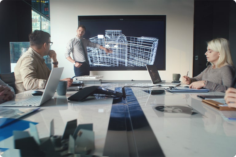 In a conference room, four people sit while a man stands center, pointing to a screen displaying a blueprint of a building.