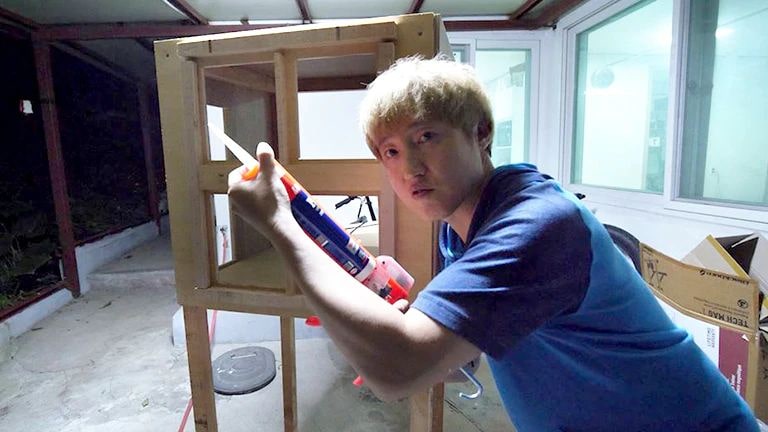 A man with yellow hair grips a silicone gun with both hands, backed by a wood-crafted object.