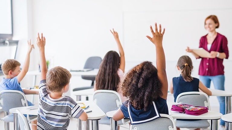 In a classroom, young kindergarten-aged children eagerly raise their hands as their teacher stands at the front.