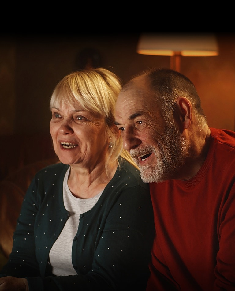 An elderly couple is sitting on a sofa, smiling and looking at the screen.