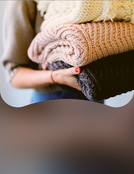 Persona che tiene in mano una pila di capi di abbigliamento lavorati a maglia e piegati, in colori neutri, tra cui maglioni color crema, beige e grigio scuro