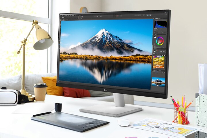 Na secretária, encontra-se um monitor ultrafino que exibe trabalho no Photoshop. Ao lado do monitor, há um candeeiro de secretária, um rato, papel e lápis de cor.