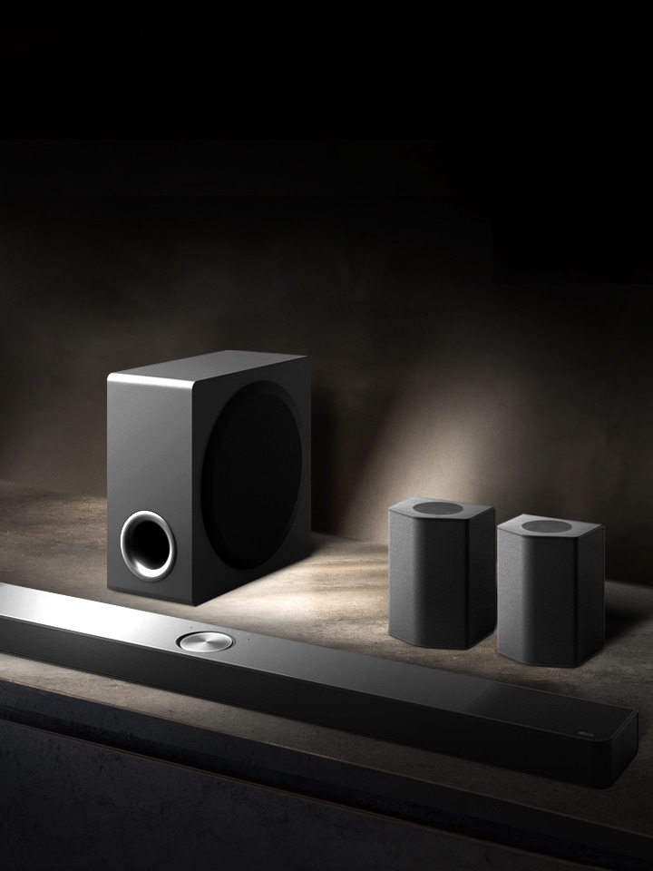 LG Soundbar, rear speakers, and subwoofer are placed within an angled perspective on a brown wooden shelf in a black room, enveloped in darkness with light only casting over the sound system.