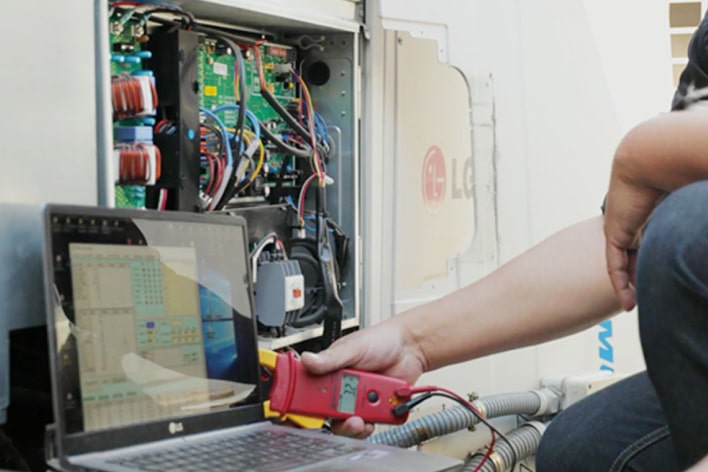 A person is using a red rectangular tool connected to a laptop, diagnosing an open LG outdoor unit, its internal wiring fully exposed.	