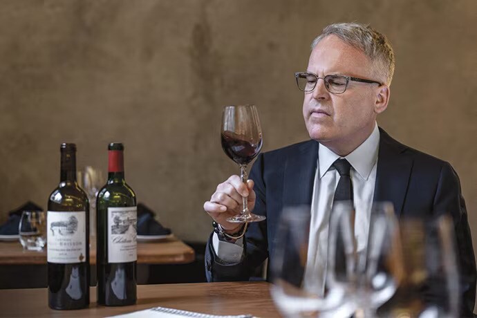 James Suckling håller ett glas vin sittande framför två flaskor vin.