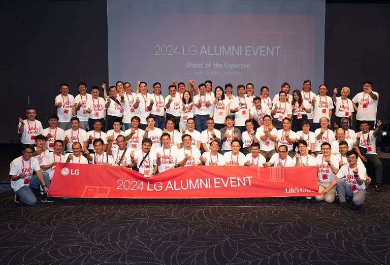 Group of people posing for a photo at the 2024 LG Alumni Event, holding a banner that reads '2024 LG Alumni Event' in front of a presentation screen.