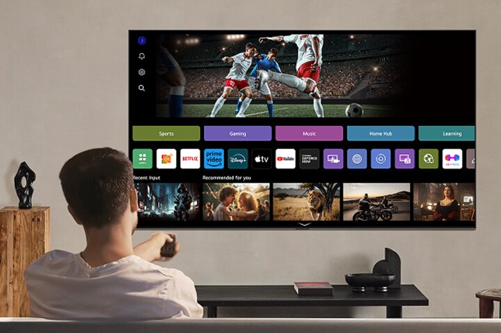 มีคนอยู่บนโซฟาถือรีโมทคอนโทรล โดยมี LG AI TV แบบติดบนผนังแสดงหน้าจอที่มี LG webOS อยู่