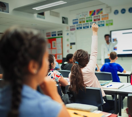 A teacher teaching through a large electronic whiteboard in an elementary school classroom and a student raising her hand to make a presentation.