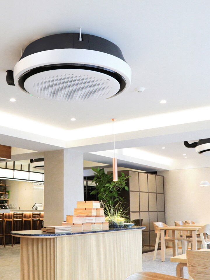 Wood and white tone dining area with tables, chairs, and kitchen displayed. LG Round Cassette mounted on the ceiling.