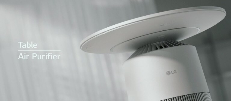 โต๊ะฟอกอากาศ LG Puricare สีขาว รูปทรงกระบอกพร้อมแผ่นวงกลมด้านบนตั้งอยู่ในห้องที่มีผ้าม่านสีขาวและแสงแดดส่องถึง