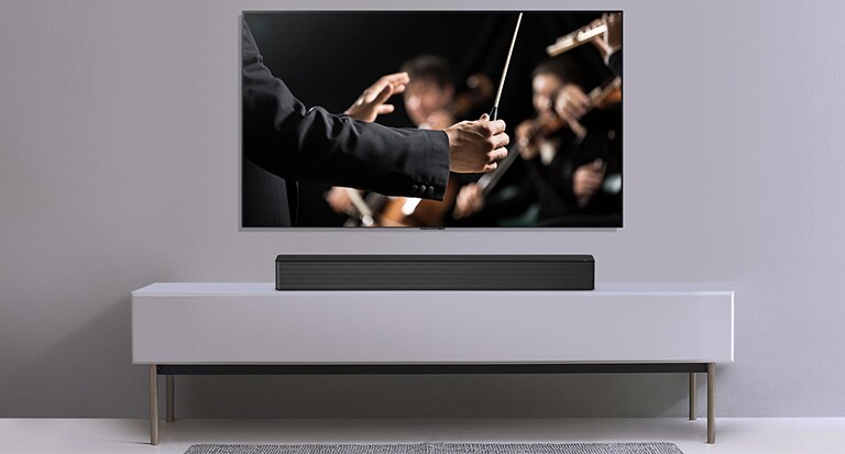 Gri bir duvara asılı TV ile altındaki gri rafta duran LG Soundbar. Orkestrayı yöneten orkestra şefini gösteren TV.
