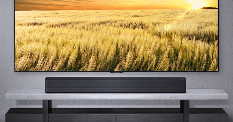 Gri bir duvara asılı TV ile altındaki gri rafta duran LG Soundbar. Rafın altında duran Blue-Ray diski.