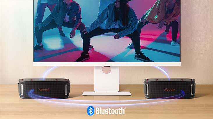 LG Smart Monitor is showing a dancing scene and two units of xboom Bounce are placed on both side of the monitor. Virtual blue light streak is connecting all 3 of them to show they're connected via bluetooth. 