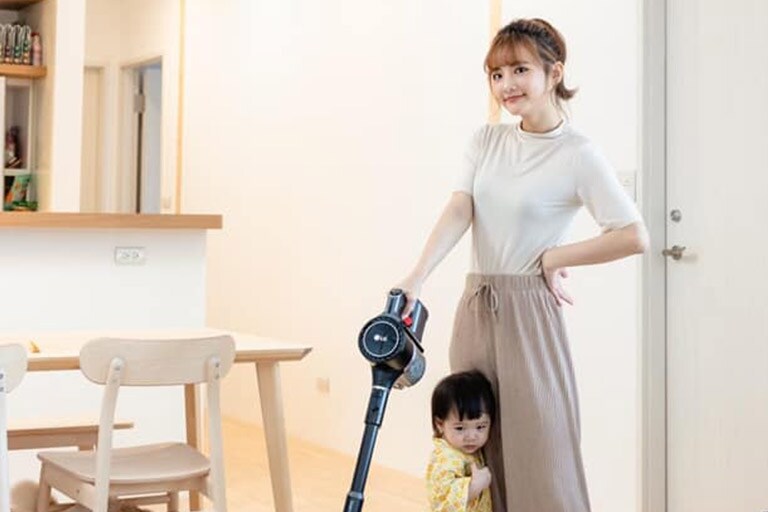 A woman stands in her kitchen easily gripping the vacuum with one hand while her child holds on to her leg.