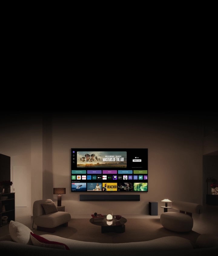 A close-up of an LG TV screen showing the buttons Home Office, Game, and Music over a banner for Masters of the Air zooms out to show the TV mounted on a wall in a living room. The following logos are displayed on the TV screen in the image: LG Channels, Netflix, Prime Video, Disney TV, Apple TV, YouTube, Spotify, Twitch, GeForce Now and Udemy.