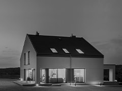 An outdoor unit located between a snow-covered brick wall and a two-storied house with wide transparent windows showing through a living room.	