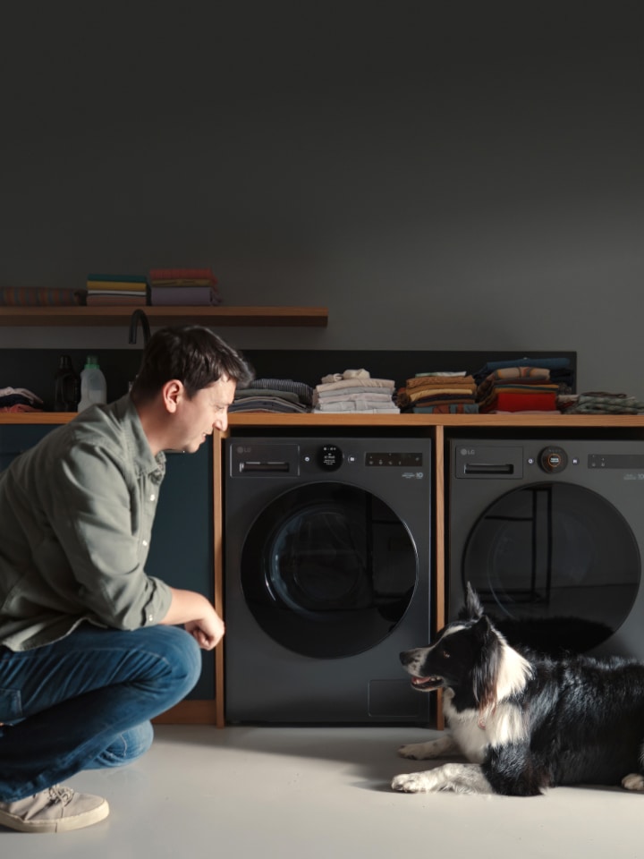 In a home laundry room, a man and a dog are facing each other, with two matte black LG washing machines visible directly behind them.