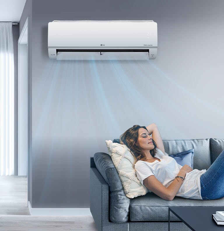 A woman relaxes on a grey sofa beneath an LG Dual Inverter air conditioner, enjoying cool air in a modern living room.