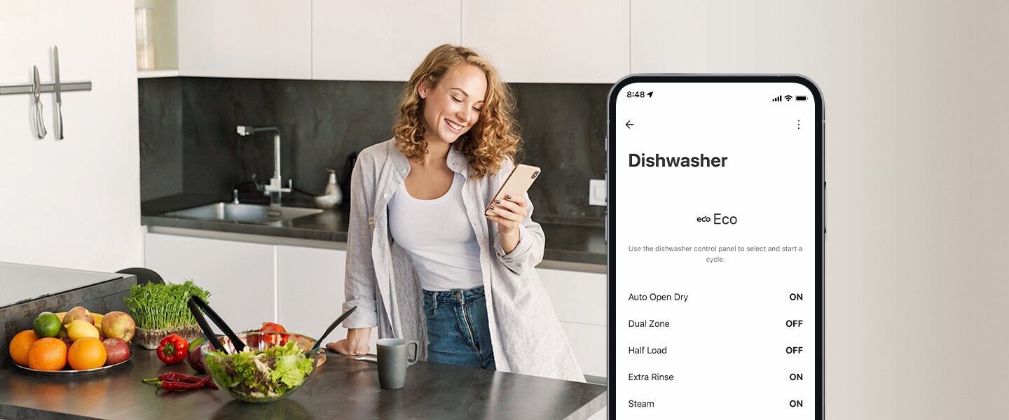 Eine Frau guckt in der Küche auf ihr Handy und auf dem Smartphone sieht man die Einstellungen in der ThinQ App. 