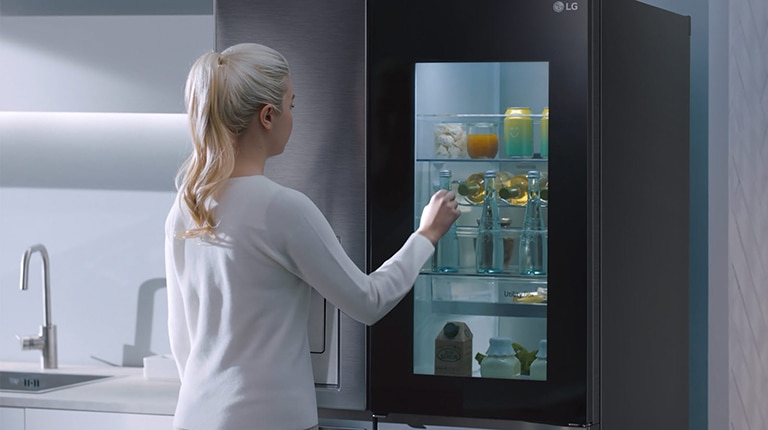 A woman opens an LG refrigerator with a see-through door in a modern kitchen, revealing drinks and groceries.