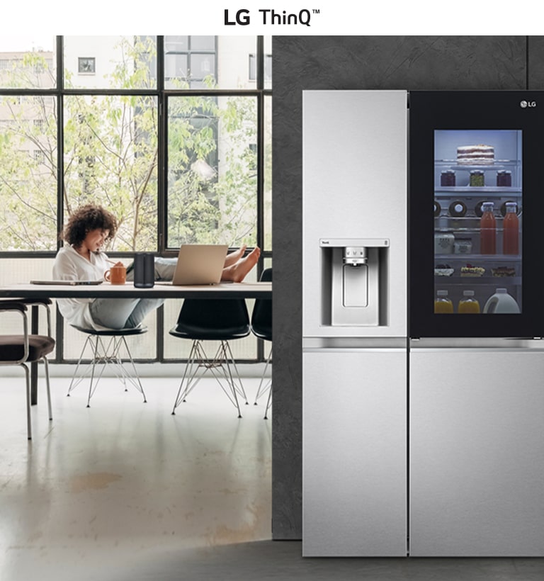 LG ThinQ refrigerator stocked with food and drinks, next to a person working on a laptop by large windows with greenery.