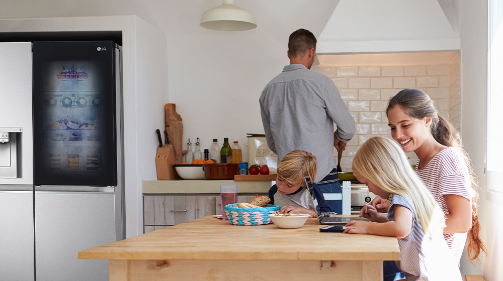 Man cooking at a counter, woman and children using devices at a wooden table, LG fridge on the left.