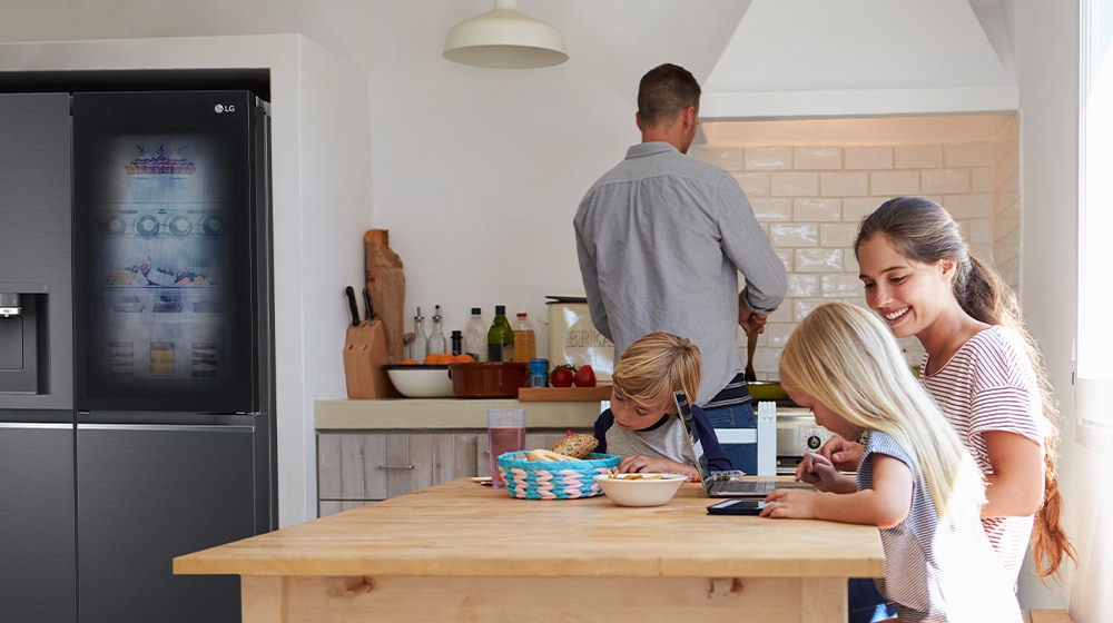 The whole family is sitting at the table preparing a meal. InstaView refrigerator installed on one side of the kitchen is creating cool air quickly.