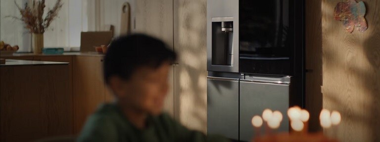 An out-of-focus child is in the centre of a kitchen. Behind him is a fridge and wooden storage drawers.