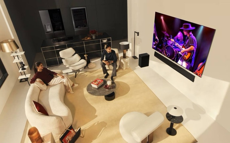 A modern living room setup with a large wall-mounted TV showcasing a live music performance. The space features luxurious seating, minimalist decor, a sleek open kitchen in the background, and a cozy atmosphere perfect for entertainment.