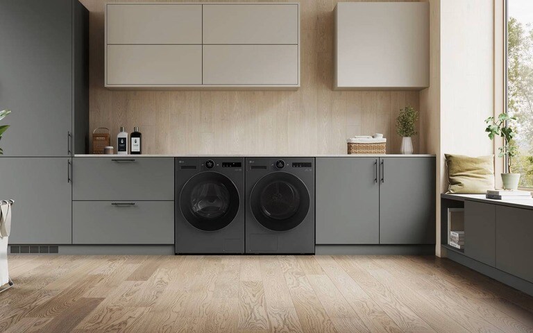 A sleek laundry room with matching black washer and dryer units built into grey cabinetry, light wood accents, and a large window bringing in natural light.