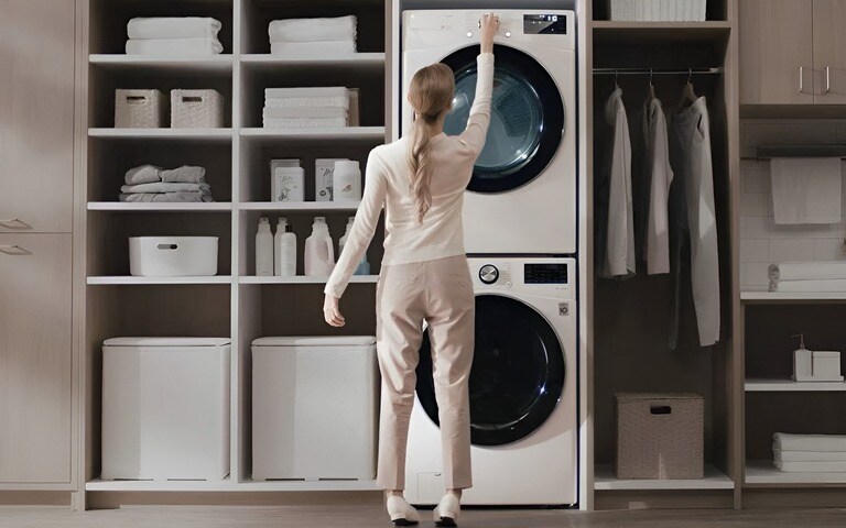 A woman adjusts settings on a stacked white washer and dryer in a well-organised laundry space with shelves holding neatly arranged laundry essentials and folded clothes.