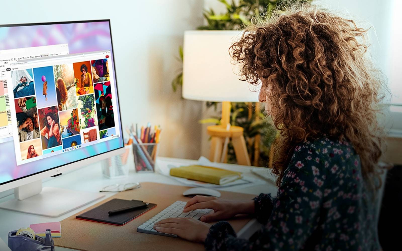 person working at an LG smart monitor, displaying images and documents. The desk setup includes a plant, lamp, and notebooks in a cosy home office environment.