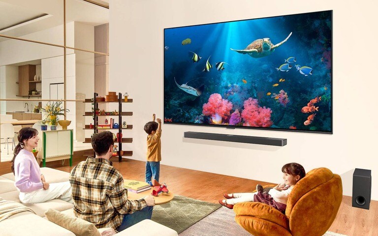 A family in a living room watching an underwater scene with vibrant coral and sea creatures on a large wall-mounted LG TV. Below the LG TV, a sleek LG soundbar and subwoofer enhance the viewing experience in the spacious and modern living area.