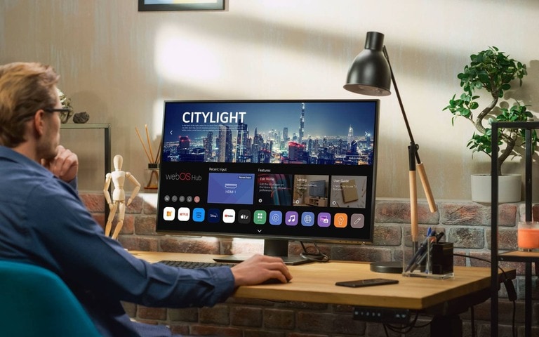 Person seated at a desk using an LG Smart Monitor displaying a LG webOS interface with various apps. The workspace includes a lamp, plants, and a modern decor with a brick wall, creating a cozy office setting.