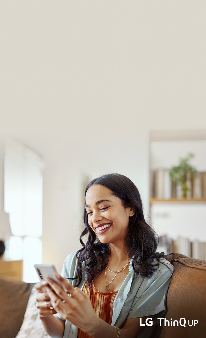 A smiling woman sitting on a couch using her smartphone in a bright living room.	