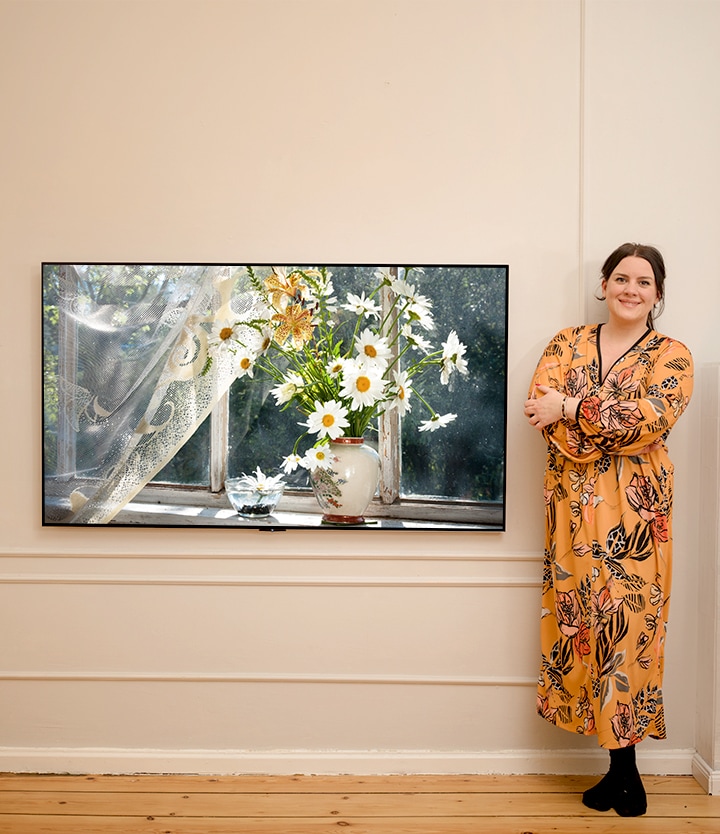The video sequentially shows the woman standing next to a TV with a vase placed by a sunlit window and smiling and the woman standing next to a washing machine.