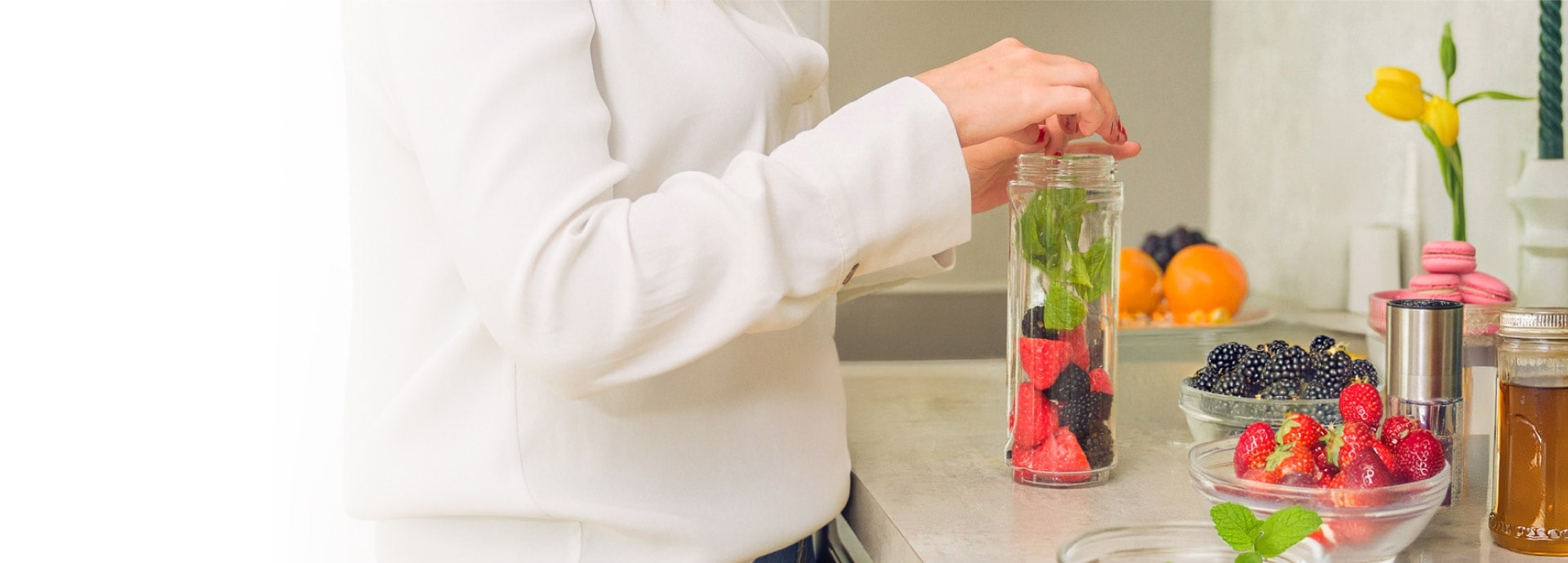 A woman is placing strawberries, blueberries, and herbs into a tall glass jar, with glass bowls filled with ingredients surrounding her.