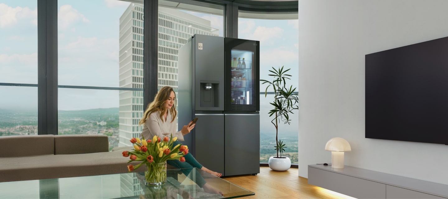 A woman uses the ThinQ app in the living room sitting on the couch which is right next to the LG InstaView American refrigerator.