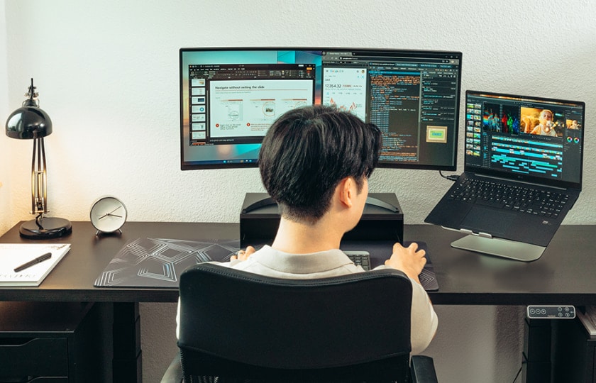 Rear view of a man at a desk with a wide monitor and laptop, showing coding and productivity apps on the screens.