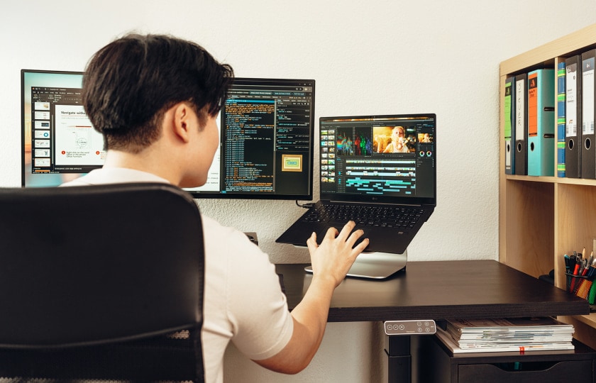 Man working in a home office with dual monitors and an elevated LG gram laptop displaying coding and video editing software, viewed from behind.