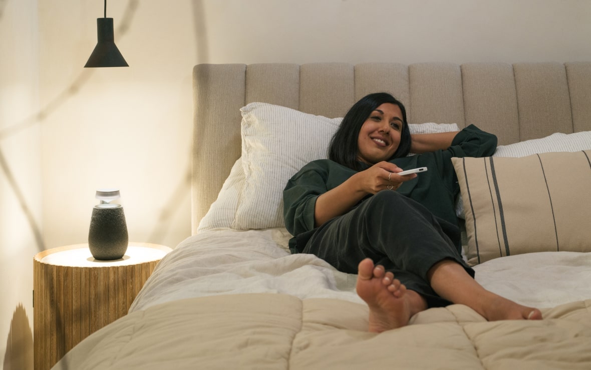 Lisa Mistry is lying in bed, using a remote control next to the LG Xboom XO2T with its Mood Lighting feature turned on.