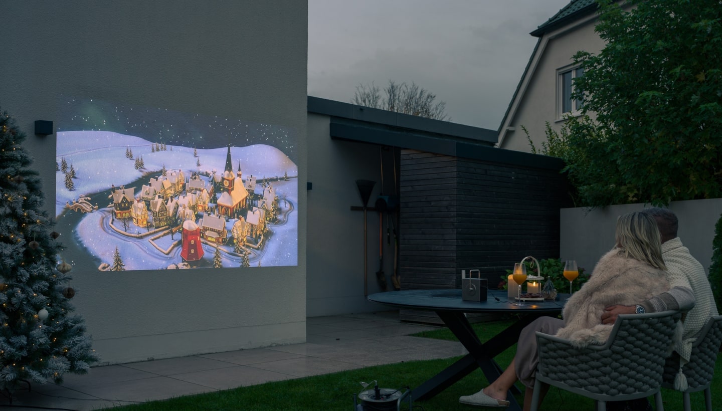 A couple enjoying an outdoor movie night with an LG CineBeam projector displaying a festive winter scene on a wall.