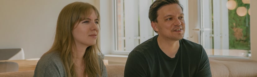 Couple sitting on a sofa playing video games together in a bright living room with large windows.