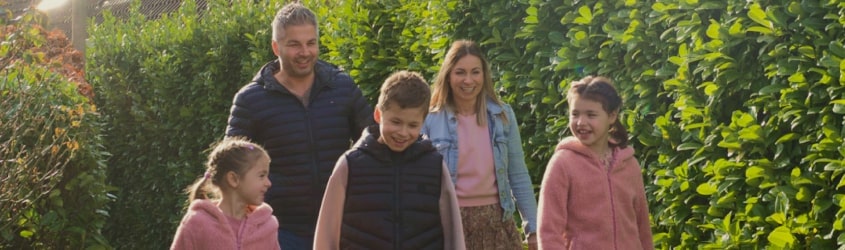 Family of five enjoying a walk outdoors, smiling and dressed in casual attire, surrounded by green hedges.