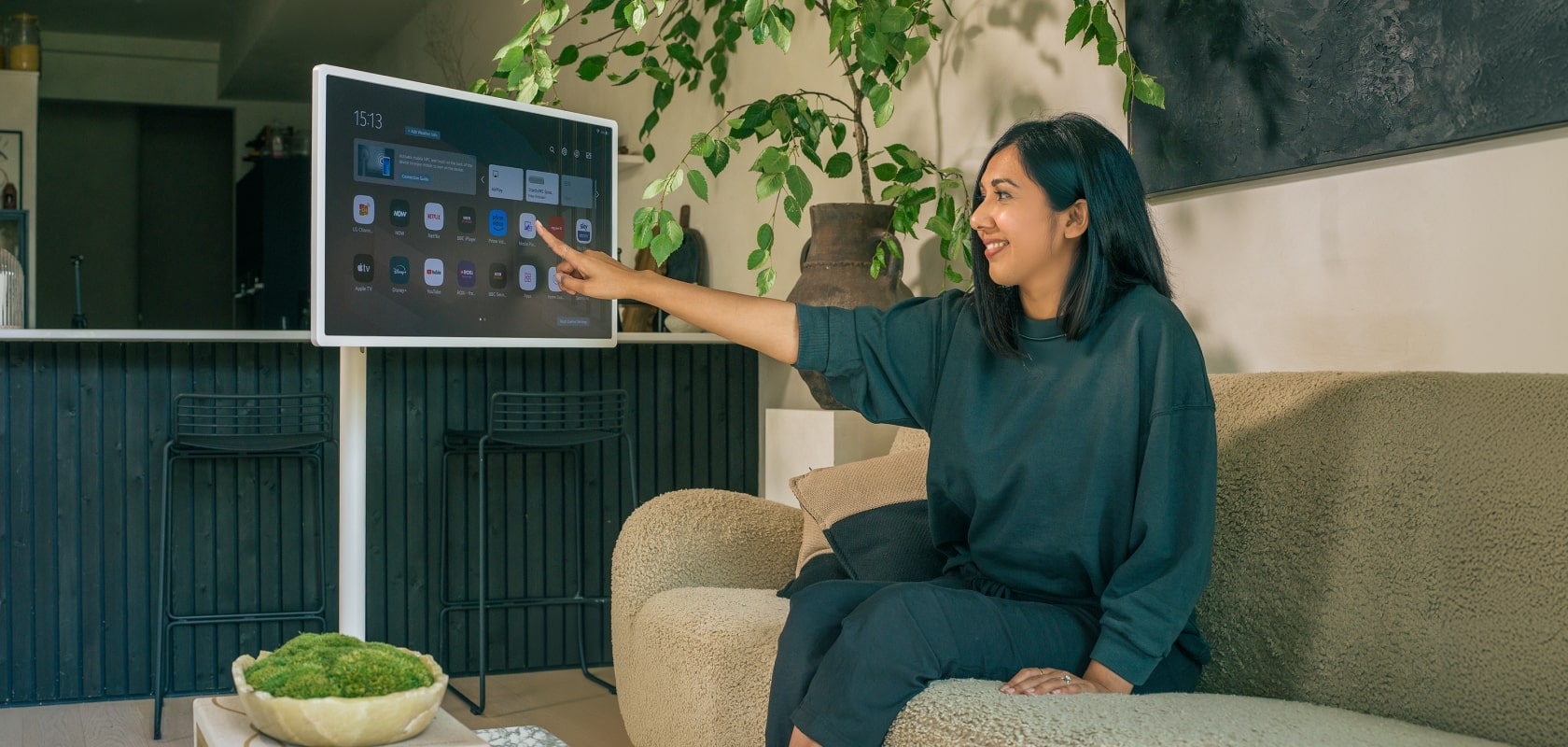 Living room with a woman interacting with an LG StanbyME screen while sitting on a comfortable sofa, surrounded by plants.