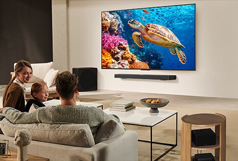 Một gia đình ngồi trên ghế sofa cùng xem chiếc TV LG treo tường phía trên LG Soundbar. Zero Connect Box đặt gọn gàng bên trong bàn trà.