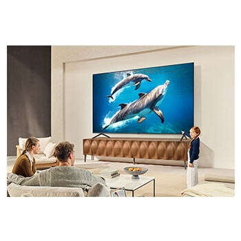 A family sits on a couch facing a LG QNED TV sitting above a TV stand with a little girl pointing to a screen depicting two dolphins. The title talks about how you can enjoy high resolution on a massive screen.